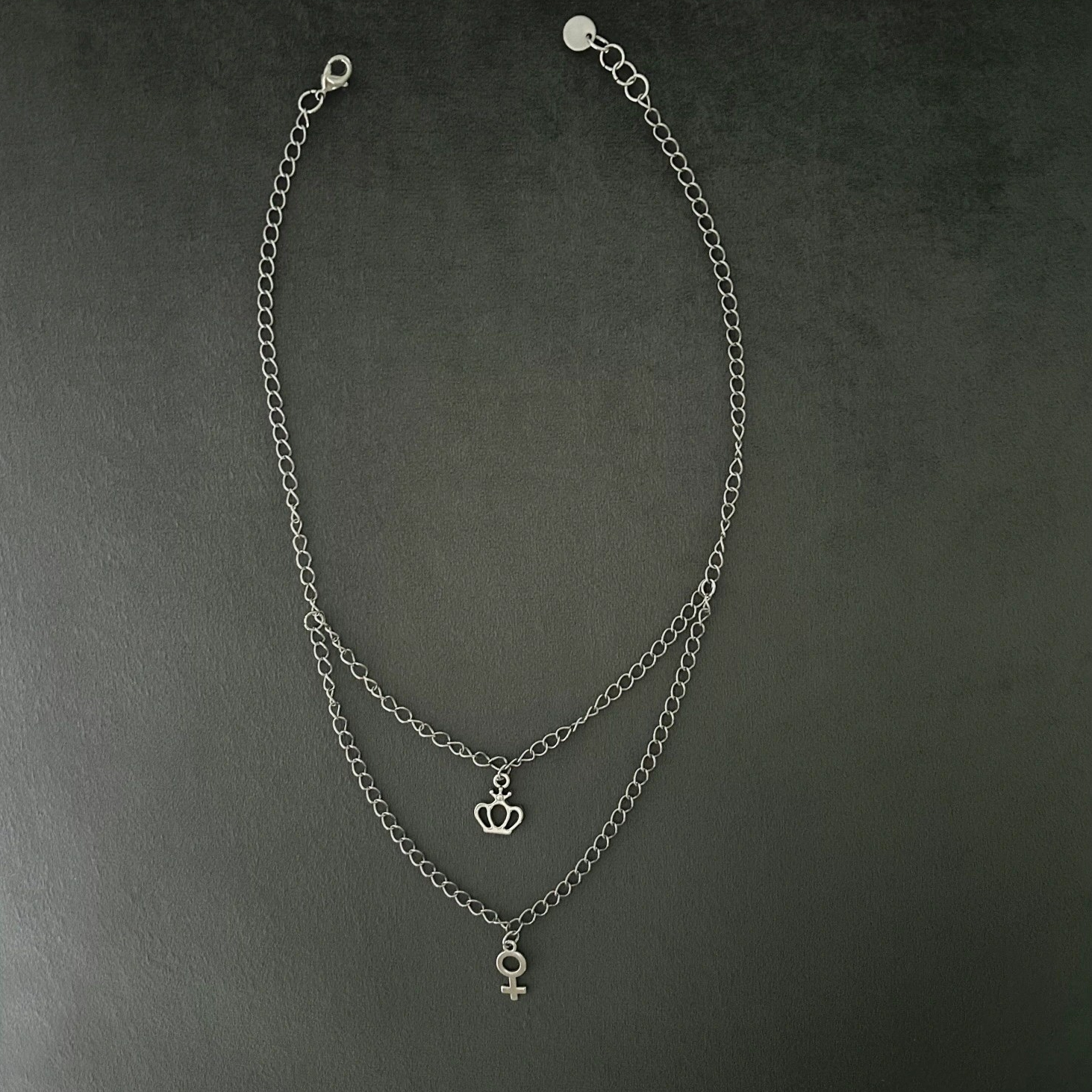 Halsband i silvernyans med dubbelkedja nedtill. Smycket förses med två dekorativa berlocker i form av kvinnosymbol och en krona.