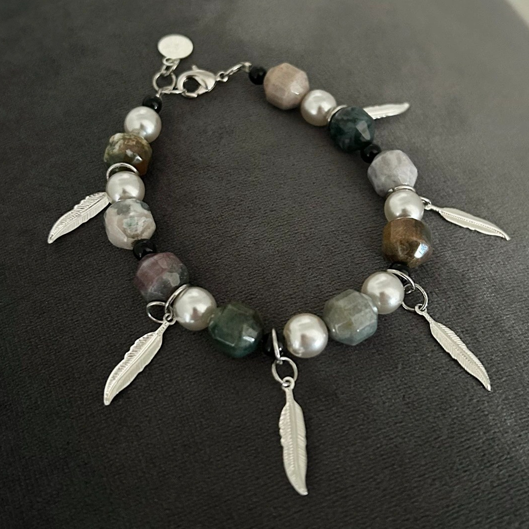 Ett dekorativt armband med Agatstenar i jordnära toner med skiftningar i grått, lila och grönt. Stenarna är något kantiga i formen. Fyra dekorativa hängen i form av fjädrar i silvriga toner tillför smycket en extra snygg detalj.