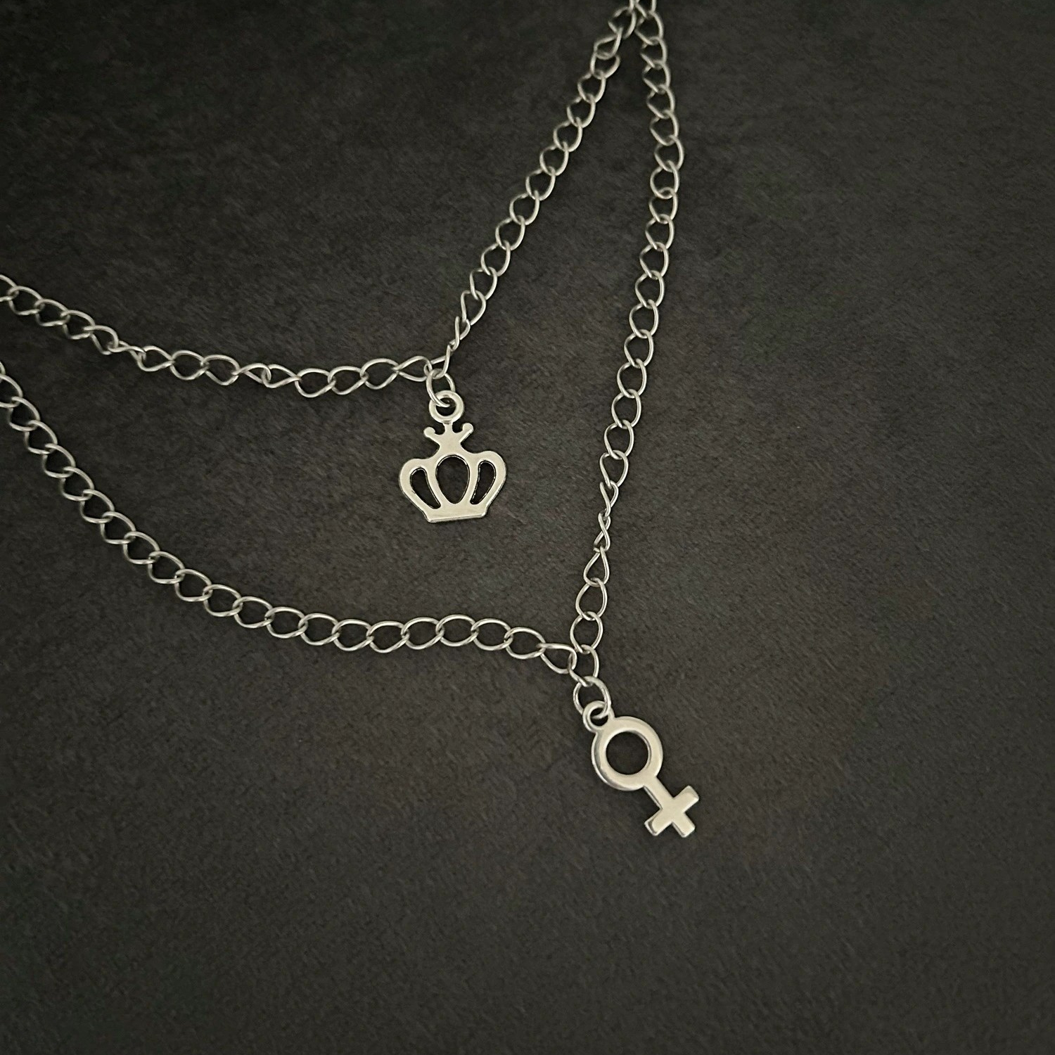 Halsband i silvernyans med dubbelkedja nedtill. Smycket förses med två dekorativa berlocker i form av kvinnosymbol och en krona.